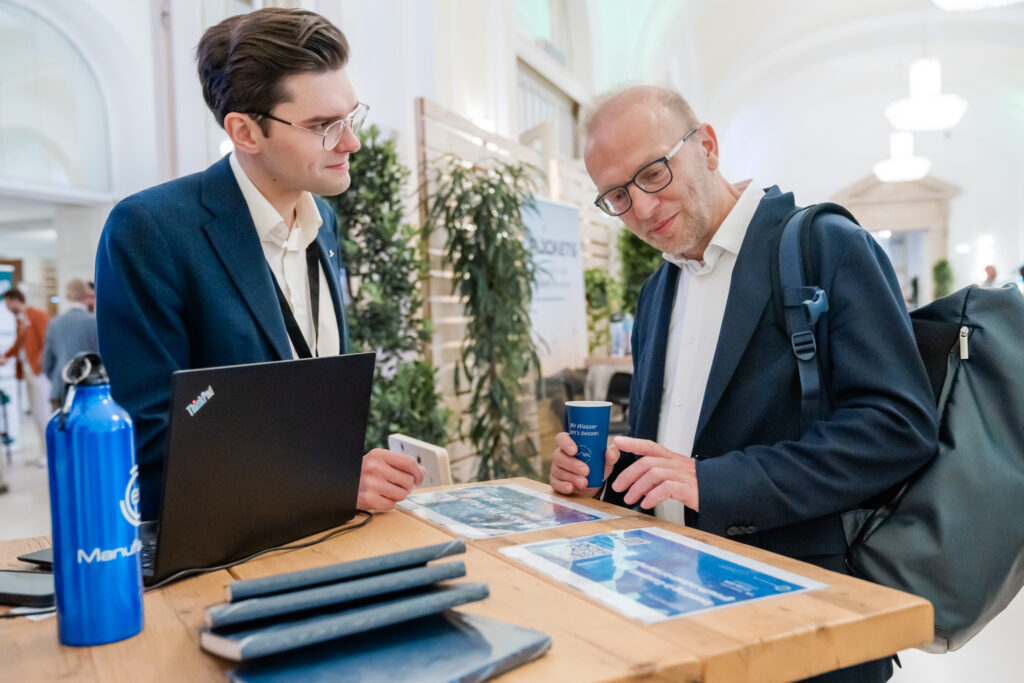 Zwei Männer in Business-Kleidung stehen an einem Holztisch während einer Networking-Veranstaltung. Der Mann links trägt einen dunkelblauen Anzug und eine Brille, steht hinter einem Laptop und hört aufmerksam zu. Der Mann rechts, ebenfalls im blauen Anzug, mit Brille und Rucksack, hält einen blauen Becher mit der Aufschrift 'Wir Wasser, ihr’s besser' und zeigt auf Informationsmaterial auf dem Tisch. Auf dem Tisch liegen außerdem blaue Notizbücher und eine blaue Trinkflasche mit der Aufschrift 'Manu...'. Im Hintergrund sind weitere Teilnehmende in Gespräche vertieft.