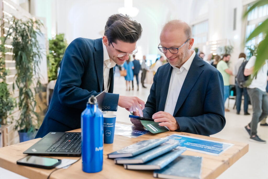 Zwei Männer in Business-Kleidung schütteln sich an einem Holztisch bei einer Networking-Veranstaltung oder Messe die Hand. Der Mann links trägt einen dunkelblauen Anzug und eine Brille und lächelt freundlich. Der Mann rechts, ebenfalls im blauen Anzug mit weißem Hemd und Rucksack, wirkt zufrieden. Auf dem Tisch liegen ein Laptop, blaue Trinkflaschen, Notizbücher und Broschüren. Im Hintergrund sind weitere Teilnehmer in Gespräche vertieft, umgeben von Pflanzen in einer hell beleuchteten, eleganten Halle.