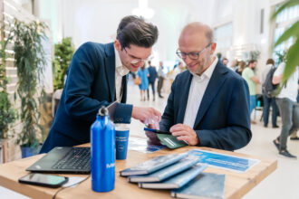 Zwei Männer in Business-Kleidung schütteln sich an einem Holztisch bei einer Networking-Veranstaltung oder Messe die Hand. Der Mann links trägt einen dunkelblauen Anzug und eine Brille und lächelt freundlich. Der Mann rechts, ebenfalls im blauen Anzug mit weißem Hemd und Rucksack, wirkt zufrieden. Auf dem Tisch liegen ein Laptop, blaue Trinkflaschen, Notizbücher und Broschüren. Im Hintergrund sind weitere Teilnehmer in Gespräche vertieft, umgeben von Pflanzen in einer hell beleuchteten, eleganten Halle.