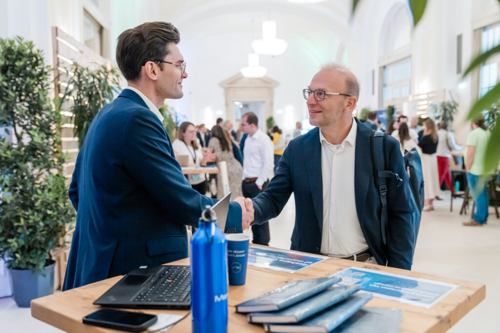 Zwei Männer in Business-Kleidung schütteln sich an einem Holztisch bei einer Networking-Veranstaltung oder Messe die Hand. Der Mann links trägt einen dunkelblauen Anzug und eine Brille und lächelt freundlich. Der Mann rechts, ebenfalls im blauen Anzug mit weißem Hemd und Rucksack, wirkt zufrieden. Auf dem Tisch liegen ein Laptop, blaue Trinkflaschen, Notizbücher und Broschüren. Im Hintergrund sind weitere Teilnehmer in Gespräche vertieft, umgeben von Pflanzen in einer hell beleuchteten, eleganten Halle.