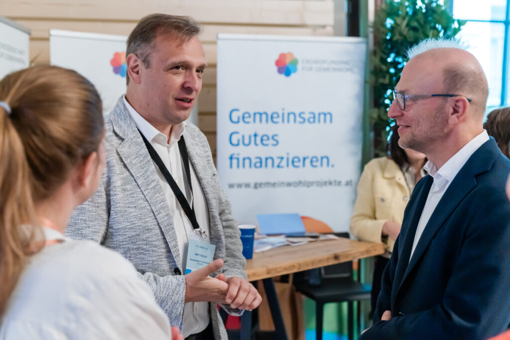 Drei Personen unterhalten sich angeregt an einem Messestand. Im Hintergrund ist ein Roll-up mit der Aufschrift „Gemeinsam Gutes finanzieren“ zu sehen, das auf Crowdfunding für Gemeinwohlprojekte hinweist. Zwei Männer, einer im grauen Sakko und der andere im dunklen Anzug, blicken sich freundlich an, während eine Frau mit zusammengebundenem Haar von hinten zu sehen ist. Auf dem Tisch liegen Informationsmaterialien und ein blauer Kaffeebecher. Pflanzen im Hintergrund verleihen der Szene eine angenehme, grüne Atmosphäre.