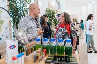 Zwei Personen stehen an einem Messestand und unterhalten sich angeregt. Der Mann links trägt ein gestreiftes Hemd, eine Brille und hält eine Hand in einer erklärenden Geste. Die Frau rechts trägt ein schwarz-weiß gestreiftes Hemd mit gelben Sternen und einen roten Schal. Auf dem Tisch vor ihnen stehen mehrere Glasflaschen mit grüner Flüssigkeit sowie kleine Fläschchen mit verschiedenen farbigen Pulvern. Im Hintergrund sind weitere Messebesucher und Stände zu sehen, die von Pflanzen dekoriert sind. Die Szene wirkt lebhaft und zeigt einen Austausch über nachhaltige Produkte.