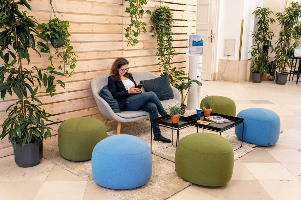 Eine Frau sitzt entspannt auf einem grauen Sessel in einer gemütlich gestalteten Lounge-Ecke mit Holzvertäfelung und vielen grünen Pflanzen. Sie trägt eine Brille, ein dunkles Sakko, Jeans und schwarze Stiefeletten und ist in ihr Smartphone vertieft. Vor ihr steht ein schwarzer Couchtisch mit kleinen Topfpflanzen, einer blauen Pappkaffeetasse und Zeitschriften. Um den Tisch sind grüne und blaue Poufs auf einem hellen Teppich arrangiert, im Hintergrund steht ein Wasserspender. Die Atmosphäre wirkt einladend und entspannt.