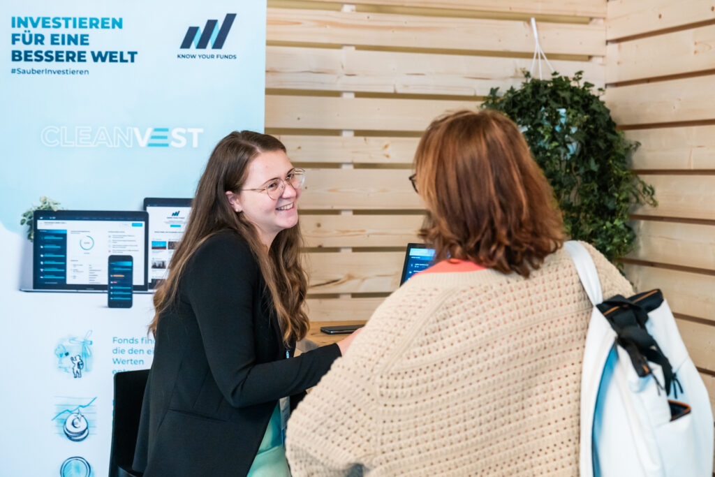 Eine junge Frau mit langen braunen Haaren, Brille und schwarzem Blazer steht an einem Messestand von „CLEANVEST“ und lächelt freundlich. Sie spricht mit einer anderen Frau, die einen beigefarbenen Strickpullover und einen weißen Rucksack trägt. Im Hintergrund ist ein Roll-up mit der Aufschrift „INVESTIEREN FÜR EINE BESSERE WELT“ sowie das Logo „KNOW YOUR FUNDS“ zu sehen. Auf dem Tisch steht ein Laptop, und die Atmosphäre wirkt freundlich und einladend.