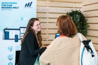 Eine junge Frau mit langen braunen Haaren, Brille und schwarzem Blazer steht an einem Messestand von „CLEANVEST“ und lächelt freundlich. Sie spricht mit einer anderen Frau, die einen beigefarbenen Strickpullover und einen weißen Rucksack trägt. Im Hintergrund ist ein Roll-up mit der Aufschrift „INVESTIEREN FÜR EINE BESSERE WELT“ sowie das Logo „KNOW YOUR FUNDS“ zu sehen. Auf dem Tisch steht ein Laptop, und die Atmosphäre wirkt freundlich und einladend.