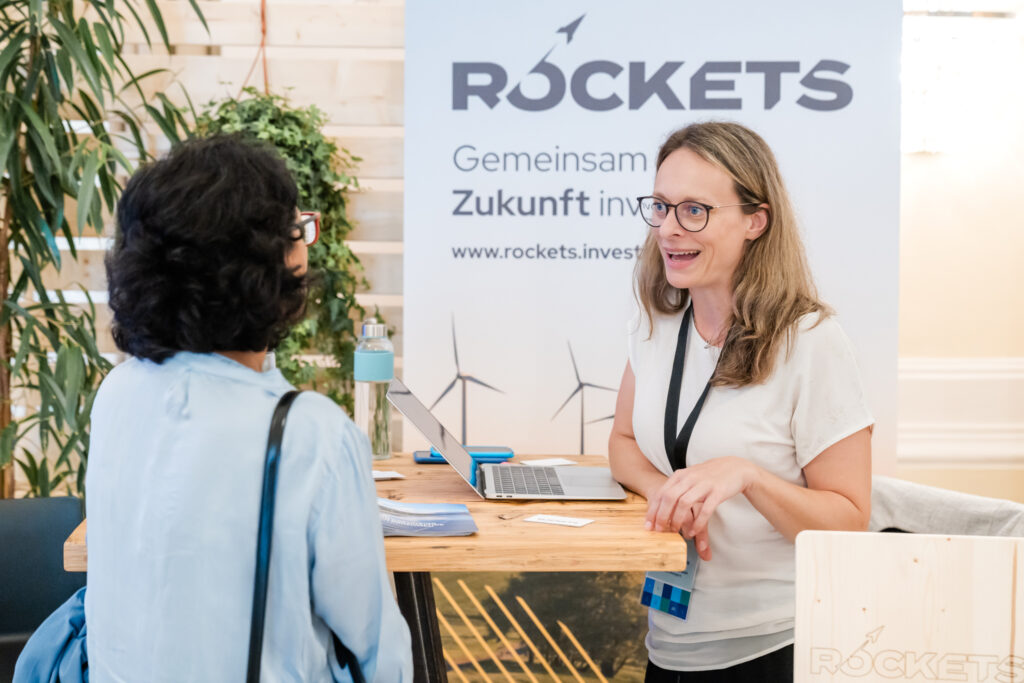 Zwei Frauen stehen an einem Messestand von „ROCKETS“. Die Frau auf der rechten Seite trägt eine weiße Bluse, eine Brille und ein Namensschildband um den Hals. Sie lächelt freundlich, während sie mit der anderen Frau spricht. Auf dem Tisch vor ihr steht ein aufgeklappter Laptop, daneben liegen Flyer und Visitenkarten. Im Hintergrund ist ein Roll-up mit der Aufschrift „ROCKETS - Gemeinsam in die Zukunft investieren“ sowie einer Abbildung von Windrädern zu sehen. Die Frau links trägt eine hellblaue Bluse und eine schwarze Tasche über der Schulter. Die Atmosphäre wirkt offen und einladend.