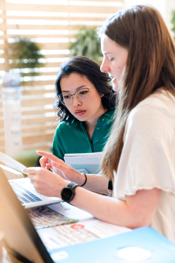 Zwei Frauen sind in ein konzentriertes Gespräch vertieft. Die Frau links trägt eine grüne Bluse und eine Brille mit dünnem Rahmen, während sie aufmerksam auf den Laptop-Bildschirm schaut. Sie hält einige Dokumente in der Hand. Die Frau rechts, in einem weißen Oberteil und mit einer schwarzen Smartwatch am Handgelenk, erklärt etwas und zeigt dabei auf den Bildschirm. Auf dem Tisch liegen verschiedene Papiere und ein blauer Ordner. Im Hintergrund sorgen Holzjalousien und einige Pflanzen für eine helle, freundliche Atmosphäre.