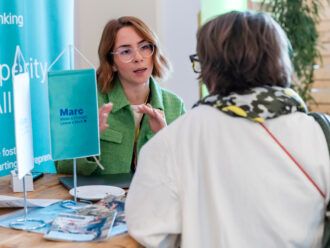 Zwei Frauen sitzen an einem Tisch und unterhalten sich konzentriert. Die Frau links trägt eine grüne Jacke und eine Brille mit rundem Rahmen, während sie auf ein Dokument zeigt. Die Frau rechts, von hinten zu sehen, trägt eine weiße Jacke und ein gemustertes Halstuch. Auf dem Tisch liegen mehrere Papiere sowie ein hellblauer Aufsteller mit der Aufschrift „Marc – Make a Change, Leave a Mark.“ Im Hintergrund ist ein blaues Roll-up-Display mit teilweise lesbarem Text und ein paar Pflanzen zu sehen. Die Szene wirkt wie ein Beratungsgespräch auf einer Messe oder Konferenz.
