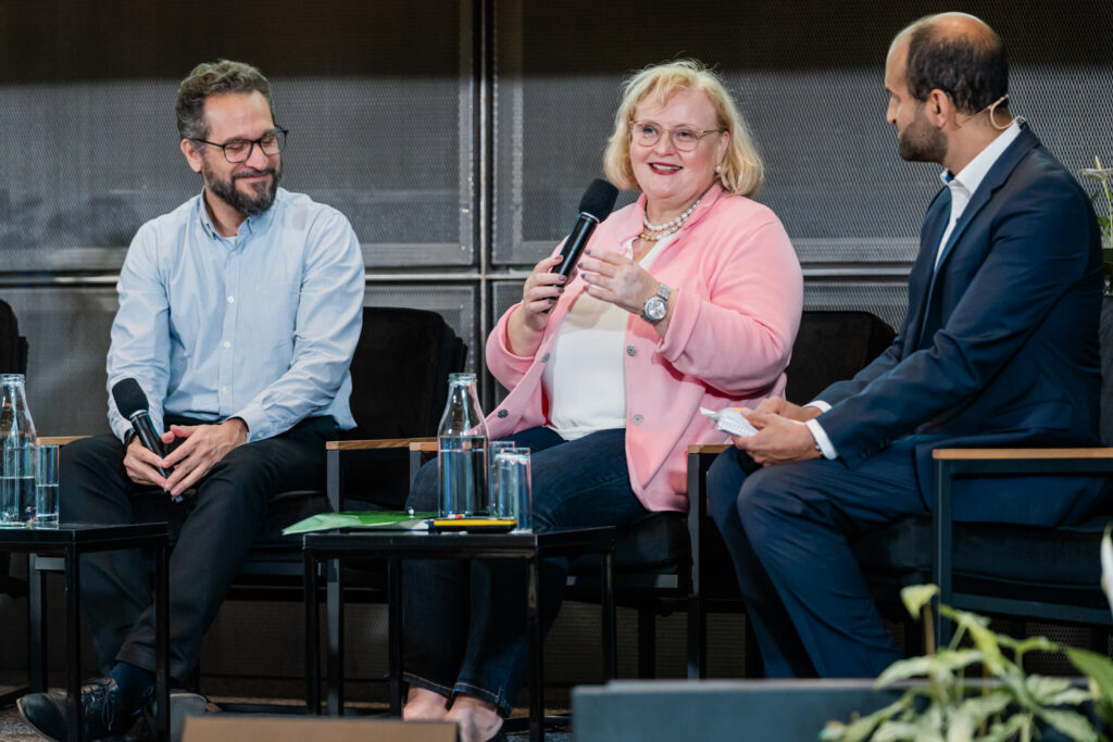 Eine Frau in einem hellrosa Blazer, weißem Top und dunkler Jeans spricht mit einem Mikrofon in der Hand, während sie freundlich lächelt. Sie trägt eine Perlenkette und eine Armbanduhr. Links von ihr sitzt ein Mann mit Brille und hellem Hemd, der sie aufmerksam und lächelnd anschaut. Rechts sitzt ein weiterer Mann in einem dunklen Anzug, der ein Notizbuch hält und ebenfalls aufmerksam zuhört. Auf dem Tisch vor ihnen stehen Wasserflaschen und Gläser. Im Hintergrund ist eine dunkle Wandverkleidung zu sehen.