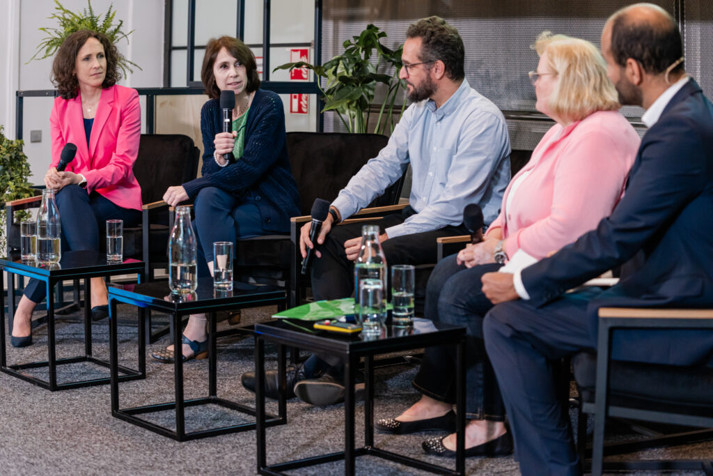 Eine Podiumsdiskussion mit fünf Teilnehmenden, die in einer Reihe auf schwarzen Stühlen sitzen. Von links nach rechts: Eine Frau in einem pinken Blazer hält ein Mikrofon und hört aufmerksam zu. Daneben spricht eine Frau mit braunen Haaren in einem dunkelblauen Cardigan und grünem Oberteil in ein Mikrofon. Ein Mann mit Brille, Bart und hellblauem Hemd hält ebenfalls ein Mikrofon und schaut die sprechende Frau an. Neben ihm sitzt eine Frau mit blondem Haar, einem rosa Blazer und einer Perlenkette, die ein Mikrofon in der Hand hält. Ganz rechts sitzt ein Mann in einem dunklen Anzug mit weißem Hemd, der Notizen macht. Auf den Tischen vor ihnen stehen Wasserflaschen, Gläser und ein grünes Notizbuch. Im Hintergrund sind Pflanzen und ein Feuerlöscher-Symbol an der Wand sichtbar.