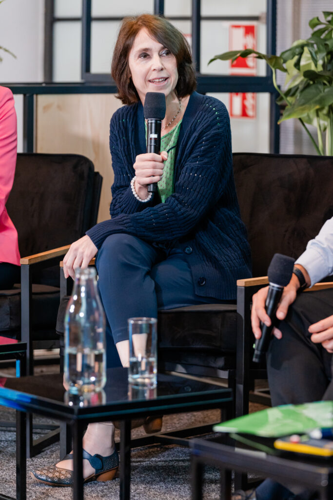 Eine Frau sitzt auf einem schwarzen Stuhl während einer Podiumsdiskussion. Sie hält ein Mikrofon in der rechten Hand und spricht, während sie leicht lächelt. Sie trägt ein grünes Oberteil mit einem dunkelblauen Cardigan und einer dunkelblauen Hose. Neben ihr ist der Arm einer weiteren Person zu sehen, die ebenfalls ein Mikrofon hält. Im Vordergrund stehen eine Wasserflasche und ein Glas Wasser auf einem kleinen schwarzen Tisch. Im Hintergrund sind Pflanzen und ein Teil einer Brandschutztür mit roten Feuerlöscher-Symbolen sichtbar.