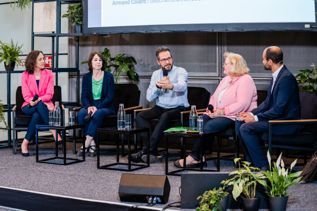 Eine Podiumsdiskussion mit fünf Teilnehmenden, die auf schwarzen Sesseln auf einer Bühne sitzen. In der Mitte spricht ein Mann mit Brille und hellblauem Hemd in ein Mikrofon, während er gestikuliert. Links von ihm sitzen zwei Frauen, eine im pinken Blazer und die andere im dunkelblauen Cardigan mit grünem Top, beide halten Mikrofone. Rechts von ihm sitzt eine Frau im rosa Blazer und ein Mann im dunklen Anzug mit Headset, der Notizen macht. Auf kleinen Tischen vor ihnen stehen Wasserflaschen und Gläser. Die Bühne ist von grünen Pflanzen umgeben.
