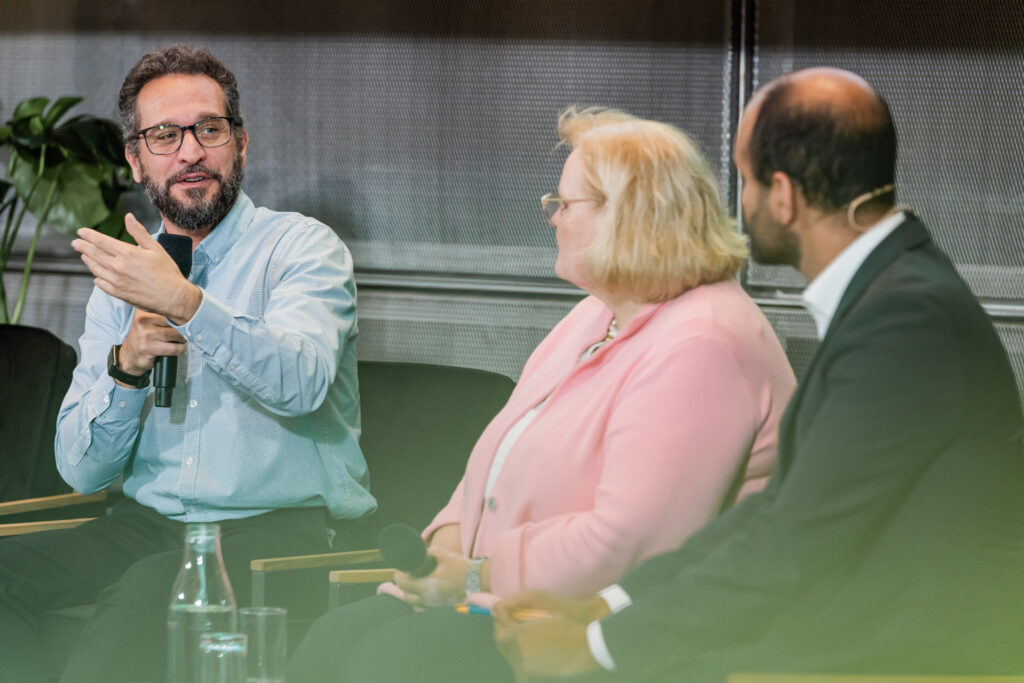 Ein Mann mit Brille, Bart und hellblauem Hemd spricht während einer Podiumsdiskussion in ein Mikrofon und gestikuliert mit der linken Hand. Neben ihm sitzen eine Frau mit blonden Haaren in einer rosa Jacke und ein Mann im dunklen Anzug mit Headset, die ihm aufmerksam zuhören. Auf dem Tisch vor ihnen stehen Wasserflaschen und Gläser. Im Hintergrund sind grüne Pflanzen und eine Wand mit Metallstruktur sichtbar.
