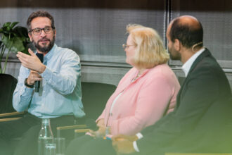 Ein Mann mit Brille, Bart und hellblauem Hemd spricht während einer Podiumsdiskussion in ein Mikrofon und gestikuliert mit der linken Hand. Neben ihm sitzen eine Frau mit blonden Haaren in einer rosa Jacke und ein Mann im dunklen Anzug mit Headset, die ihm aufmerksam zuhören. Auf dem Tisch vor ihnen stehen Wasserflaschen und Gläser. Im Hintergrund sind grüne Pflanzen und eine Wand mit Metallstruktur sichtbar.