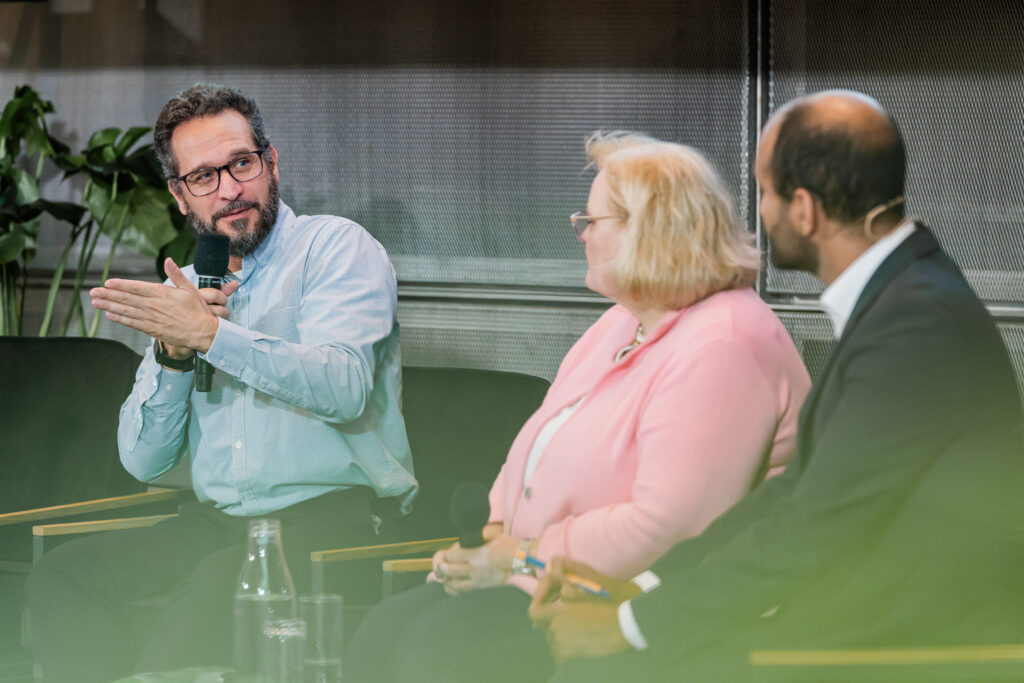 Ein Mann mit Brille, Bart und hellblauem Hemd spricht während einer Podiumsdiskussion in ein Mikrofon und gestikuliert mit der linken Hand. Neben ihm sitzen eine Frau mit blonden Haaren in einer rosa Jacke und ein Mann im dunklen Anzug mit Headset, die ihm aufmerksam zuhören. Auf dem Tisch vor ihnen stehen Wasserflaschen und Gläser. Im Hintergrund sind grüne Pflanzen und eine Wand mit Metallstruktur sichtbar.