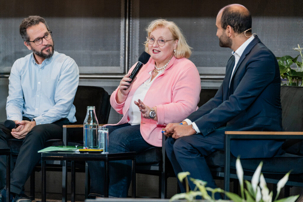Eine Nahaufnahme einer Podiumsdiskussion mit drei Teilnehmenden. In der Mitte spricht eine Frau mit blonden Haaren, Brille und rosa Jacke, während sie ein Mikrofon hält. Sie trägt eine Perlenkette und gestikuliert mit der linken Hand. Links von ihr sitzt ein Mann mit Brille und Bart in einem hellblauen Hemd, der aufmerksam zuhört. Rechts von ihr sitzt ein weiterer Mann im dunklen Anzug mit weißem Hemd, der einen Stift in der Hand hält und ebenfalls zuhört. Auf dem Tisch vor ihnen stehen Wasserflaschen und Gläser. Im Hintergrund sind grüne Pflanzen zu sehen.