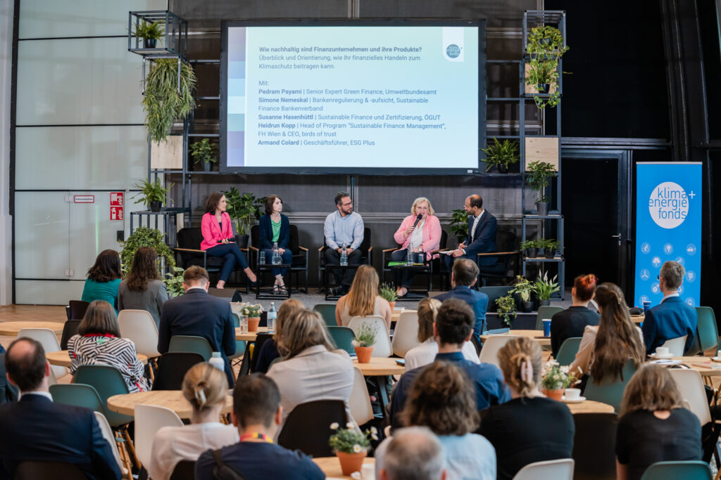 Eine Podiumsdiskussion mit fünf Teilnehmenden (drei Frauen, zwei Männer) auf einer Bühne, die von zahlreichen grünen Pflanzen umgeben ist. Im Hintergrund ist eine große Leinwand mit der Frage „Wie nachhaltig sind Finanzunternehmen und ihre Produkte?“ zu sehen. Darunter stehen die Namen und Titel der Diskutierenden: Pedram Payami, Simone Nemeskal, Susanne Hasenhüttl, Heidrun Kopp und Armand Colard. Das Publikum sitzt an runden Tischen mit kleinen Blumentöpfen. Auf der rechten Seite steht ein blaues Banner mit dem Logo von „Klima + Energie Fonds“. Die Atmosphäre wirkt professionell und fokussiert auf nachhaltige Finanzthemen.