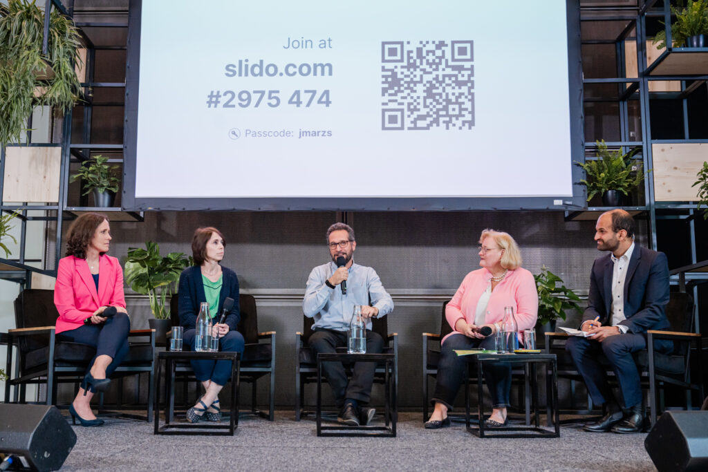 Eine Podiumsdiskussion mit fünf Teilnehmenden, die auf schwarzen Sesseln vor einer großen Leinwand sitzen. Auf der Leinwand ist ein QR-Code sowie der Hinweis „Join at slido.com #2975 474“ und ein Passcode „jmarzs“ zu sehen. Die Teilnehmenden halten Mikrofone, diskutieren angeregt und sind von mehreren grünen Pflanzen umgeben, was dem Setting eine frische Atmosphäre verleiht. Die Veranstaltung wirkt professionell, mit Fokus auf Interaktion durch das Online-Tool Slido.