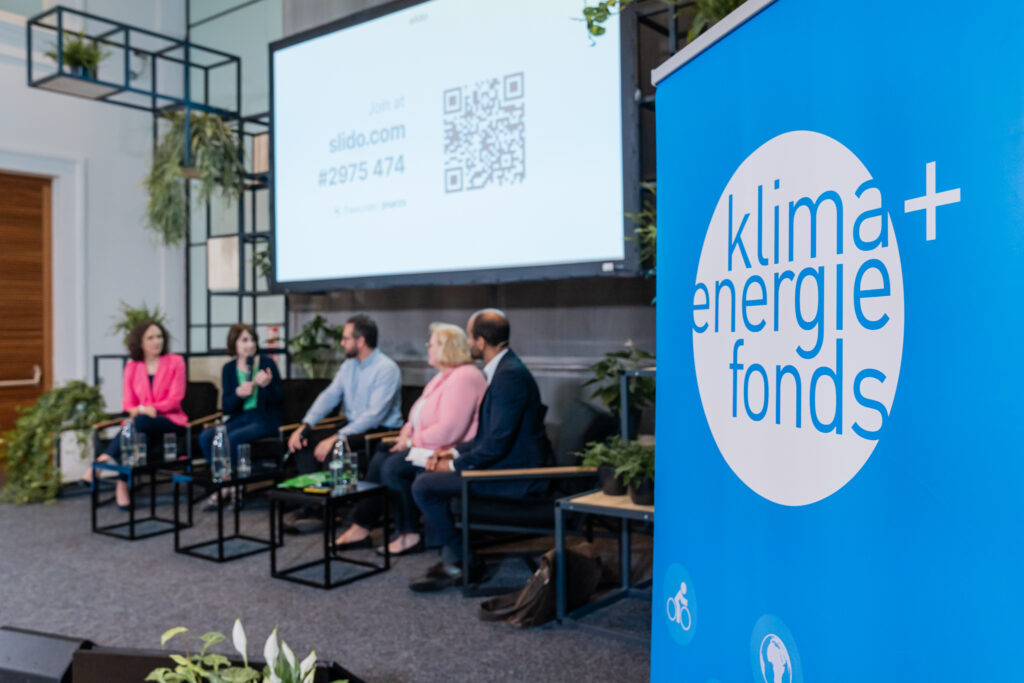 Eine Podiumsdiskussion mit fünf Teilnehmenden, die auf einer Bühne sitzen. Im Vordergrund ist ein blaues Roll-up mit dem Logo des 