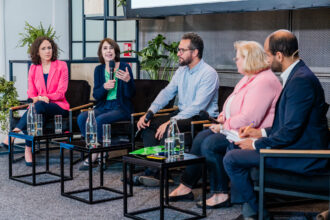 Eine Podiumsdiskussion mit fünf Personen, die auf schwarzen Sesseln auf einer Bühne sitzen. Die zweite Person von links, eine Frau in einem grünen Top und dunkelblauer Strickjacke, spricht in ein Mikrofon und gestikuliert mit der Hand. Die anderen Teilnehmenden hören aufmerksam zu. Auf den kleinen Tischen vor ihnen stehen Glasflaschen mit Wasser und Gläser. Im Hintergrund sind grüne Pflanzen und Regale zu sehen, die dem Raum eine moderne und freundliche Atmosphäre verleihen.