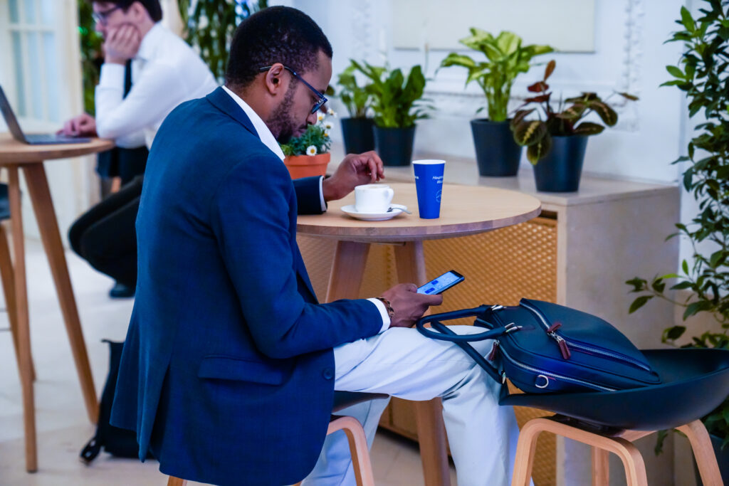 Ein Mann in einem blauen Anzug sitzt an einem runden Holztisch in einer modernen Lounge-Umgebung. Er hält ein Smartphone in der rechten Hand und blickt konzentriert darauf, während er mit der linken Hand eine Tasse Kaffee anhebt. Neben ihm auf einem Hocker liegt eine dunkelblaue Aktentasche. Im Hintergrund arbeitet eine weitere Person an einem Laptop. Der Raum ist mit mehreren grünen Topfpflanzen dekoriert, was eine angenehme, entspannte Atmosphäre schafft. Auf dem Tisch stehen zusätzlich ein blauer Pappbecher und ein kleiner Blumentopf mit weißen Gänseblümchen.