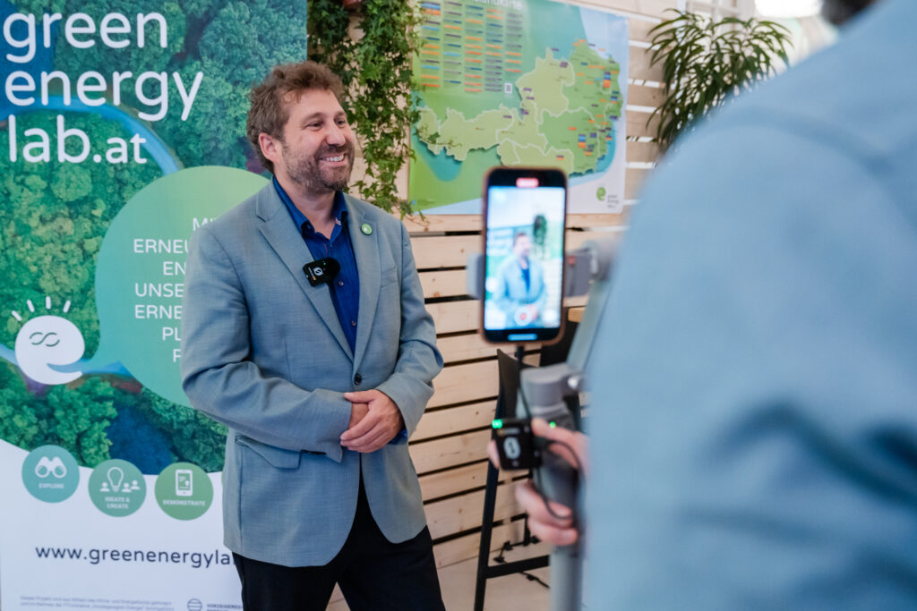 Ein Mann in einem hellblauen Sakko und dunkelblauen Hemd lächelt, während er in einem Interview gefilmt wird. Er trägt ein Ansteckmikrofon am Revers. Im Vordergrund hält eine Person ein Smartphone auf einem Gimbal, das den Mann aufnimmt. Im Hintergrund ist ein Banner von „greenenergylab.at“ mit einem grünen Waldmotiv sowie eine Karte mit bunten Markierungen zu sehen. Die Szene spielt in einem hellen Raum mit Holzwänden und grünen Pflanzen.