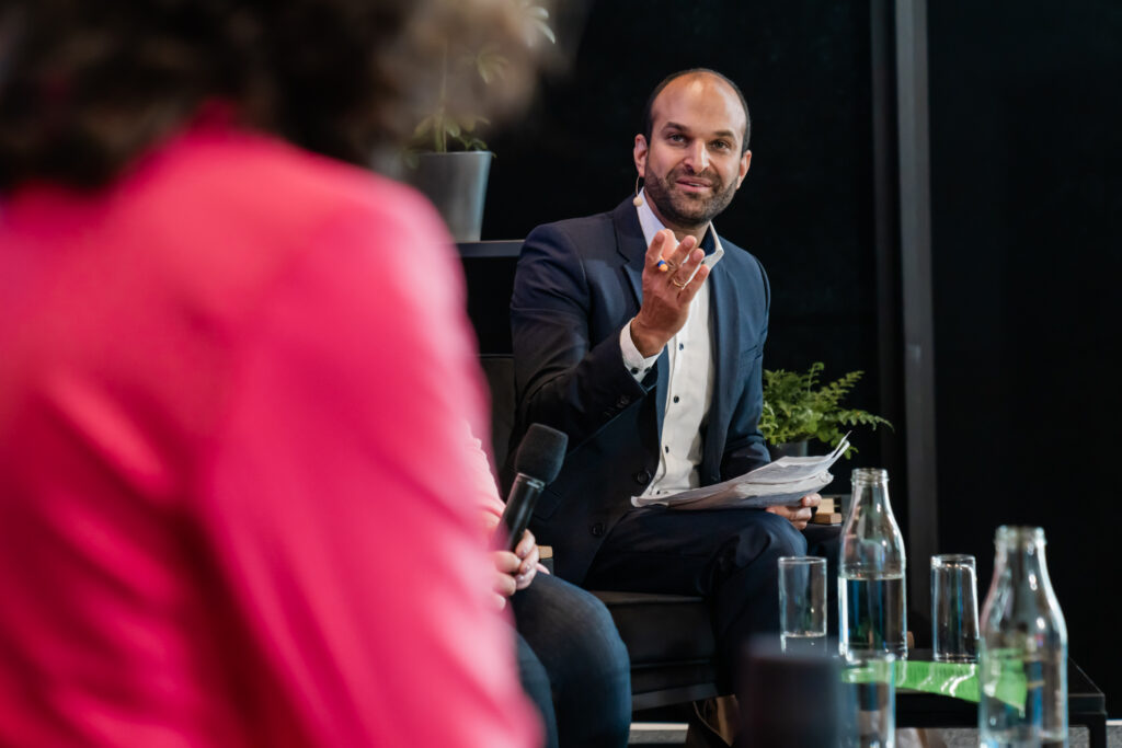 Ein Mann in einem dunkelblauen Anzug mit weißem Hemd spricht engagiert in einer Podiumsdiskussion. Er hält einige Notizzettel in der linken Hand und gestikuliert mit der rechten. Er trägt ein Headset-Mikrofon. Im Vordergrund ist unscharf eine Person in einem pinken Blazer zu sehen, die vermutlich Teil der Diskussion ist und ein Mikrofon hält. Auf dem Tisch vor ihnen stehen mehrere Glasflaschen mit Wasser und Gläser. Im Hintergrund sind einige grüne Pflanzen zu sehen.