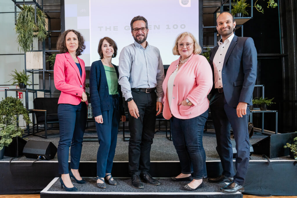 Fünf Personen posieren gemeinsam für ein Gruppenfoto auf einer Bühne, die mit Pflanzen dekoriert ist. Im Hintergrund ist ein großer Bildschirm zu sehen, auf dem „THE GREEN 100“ steht. Die Gruppe besteht aus drei Frauen und zwei Männern, die alle geschäftlich gekleidet sind. Die Frau ganz links trägt einen pinken Blazer und dunkelblaue Hose, daneben steht eine Frau in einem grünen Top mit einer dunkelblauen Strickjacke. In der Mitte befindet sich ein Mann mit Brille in einem hellblauen Hemd. Die Frau neben ihm trägt eine helle, rosa Strickjacke und Perlenkette. Ganz rechts steht ein Mann im dunkelblauen Anzug. Alle blicken lächelnd in die Kamera.