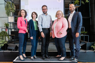 Fünf Personen posieren gemeinsam für ein Gruppenfoto auf einer Bühne, die mit Pflanzen dekoriert ist. Im Hintergrund ist ein großer Bildschirm zu sehen, auf dem „THE GREEN 100“ steht. Die Gruppe besteht aus drei Frauen und zwei Männern, die alle geschäftlich gekleidet sind. Die Frau ganz links trägt einen pinken Blazer und dunkelblaue Hose, daneben steht eine Frau in einem grünen Top mit einer dunkelblauen Strickjacke. In der Mitte befindet sich ein Mann mit Brille in einem hellblauen Hemd. Die Frau neben ihm trägt eine helle, rosa Strickjacke und Perlenkette. Ganz rechts steht ein Mann im dunkelblauen Anzug. Alle blicken lächelnd in die Kamera.
