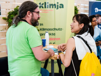 Zwei Personen stehen an einem hohen Tisch und unterhalten sich angeregt. Der Mann trägt ein hellgrünes T-Shirt und eine Brille, seine langen Haare sind zu einem Zopf gebunden. Die Frau ihm gegenüber hat dunkle Haare, trägt ein weißes Sweatshirt über den Schultern und einen gelben Turnbeutel. Im Hintergrund ist ein gelbes Banner mit der Aufschrift „klimja“ sowie ein Text zu Klimaschutzprojekten sichtbar. Der Tisch ist mit einem blauen Pappbecher, einer weißen Trinkflasche und Infomaterialien bedeckt.