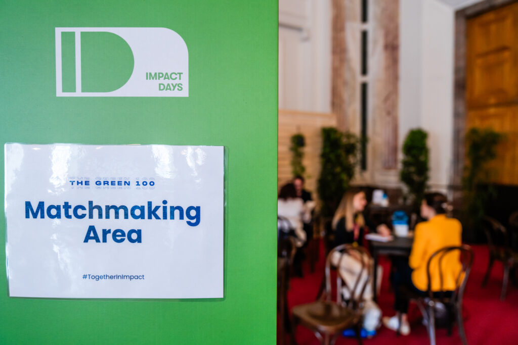 Ein Schild mit der Aufschrift „THE GREEN 100 Matchmaking Area“ und dem Hashtag #TogetherInImpact ist auf einer grünen Trennwand angebracht. Darüber befindet sich das Logo von „IMPACT DAYS“. Im unscharfen Hintergrund sitzen mehrere Personen an kleinen Tischen, offenbar in Gespräche vertieft, in einem Raum mit roten Teppichboden, hohen Fenstern und dekorativen Pflanzen. Die Szene vermittelt den Eindruck eines Networking-Bereichs auf einer Konferenz oder Veranstaltung.