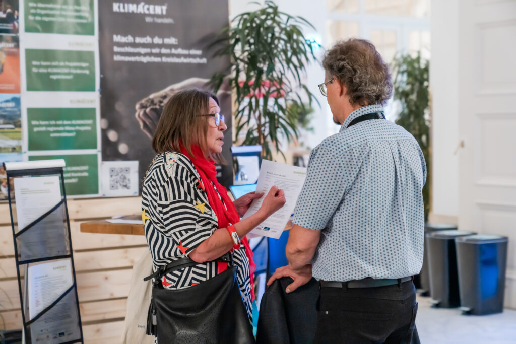 Zwei Personen stehen in einem Gespräch an einem Messestand. Die Frau links trägt eine schwarz-weiß gestreifte Bluse mit bunten Sternenmustern, einen roten Schal und eine schwarze Tasche über der Schulter. Sie hält ein Informationsblatt in der Hand und erklärt dem Mann gegenüber etwas, während sie auf das Blatt zeigt. Der Mann trägt ein hellblaues, gemustertes Hemd und eine Brille. Im Hintergrund ist ein Roll-up mit dem Schriftzug „KLIMÁCENT“ zu sehen, sowie grüne Pflanzen, Broschüren und ein moderner, heller Ausstellungsbereich.