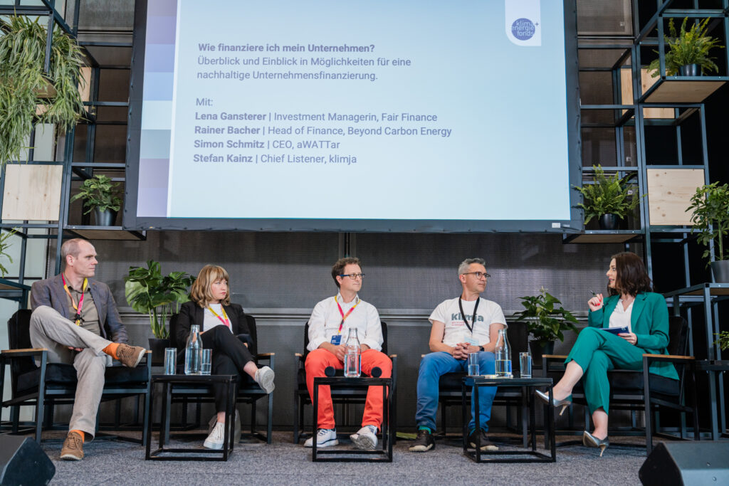 Fünf Personen sitzen auf einer Bühne in einer Podiumsdiskussion. Über ihnen ist eine große Präsentationsfolie mit dem Titel „Wie finanziere ich mein Unternehmen?“ zu sehen, die die Namen der Diskussionsteilnehmer:innen auflistet: Lena Gansterer, Rainer Bacher, Simon Schmitz und Stefan Kainz. Die Teilnehmenden sind in angeregter Diskussion, wobei eine Frau im grünen Anzug ein Mikrofon hält und spricht. Die Bühne ist von Pflanzen dekoriert, was eine lebendige Atmosphäre schafft. Vor der Bühne sitzen Zuhörer:innen, die der Diskussion aufmerksam folgen.