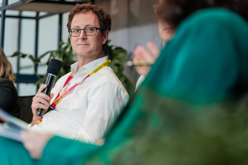 Ein Mann in einem weißen Hemd mit einem bunten Konferenz-Lanyard hält ein Mikrofon in der Hand. Er trägt eine Brille und blickt aufmerksam nach vorne, vermutlich zu einem anderen Diskussionsteilnehmenden. Im Vordergrund ist unscharf eine Person in einem grünen Anzug zu sehen, die sich im Gespräch mit dem Mann zu befinden scheint. Im Hintergrund sind grüne Pflanzen sichtbar, die der Szene eine frische Atmosphäre verleihen.