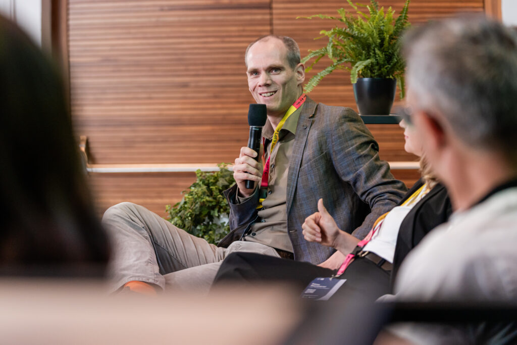 Ein Mann in einem karierten Sakko und grauer Hose spricht auf einer Podiumsdiskussion in ein Mikrofon. Er trägt ein rotes Konferenz-Lanyard um den Hals und lächelt, während er sich an das Publikum oder die anderen Diskussionsteilnehmenden wendet. Im Hintergrund sind grüne Pflanzen zu sehen, die der Veranstaltung eine natürliche Atmosphäre verleihen.
