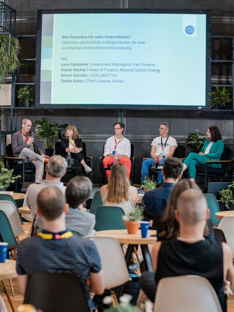 Podiumsdiskussion mit fünf Teilnehmenden, die auf einer Bühne vor einem großen Bildschirm sitzen. Der Bildschirm zeigt das Thema „Wie finanziere ich mein Unternehmen?“ mit den Namen der Diskussionsteilnehmenden: Lena Gansterer, Rainer Bacher, Simon Schmitz und Stefan Kainz. Im Publikum sitzen mehrere Zuhörer an Tischen mit kleinen Pflanzendekorationen. Die Bühne ist von grünen Pflanzen umgeben, was eine nachhaltige Atmosphäre betont.