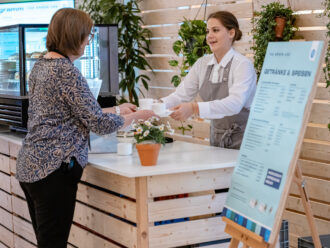Eine Barista in weißem Hemd und grauer Schürze reicht einer Kundin eine Tasse Kaffee über den Tresen eines modernen Café-Standes. Der Tresen besteht aus hellem Holz, dekoriert mit kleinen Blumentöpfen. Im Hintergrund sind grüne Hängepflanzen zu sehen. Rechts im Bild steht eine große Speisekarte mit der Überschrift „GETRÄNKE & SPEISEN“. Links befindet sich eine Glasvitrine mit Gebäck.