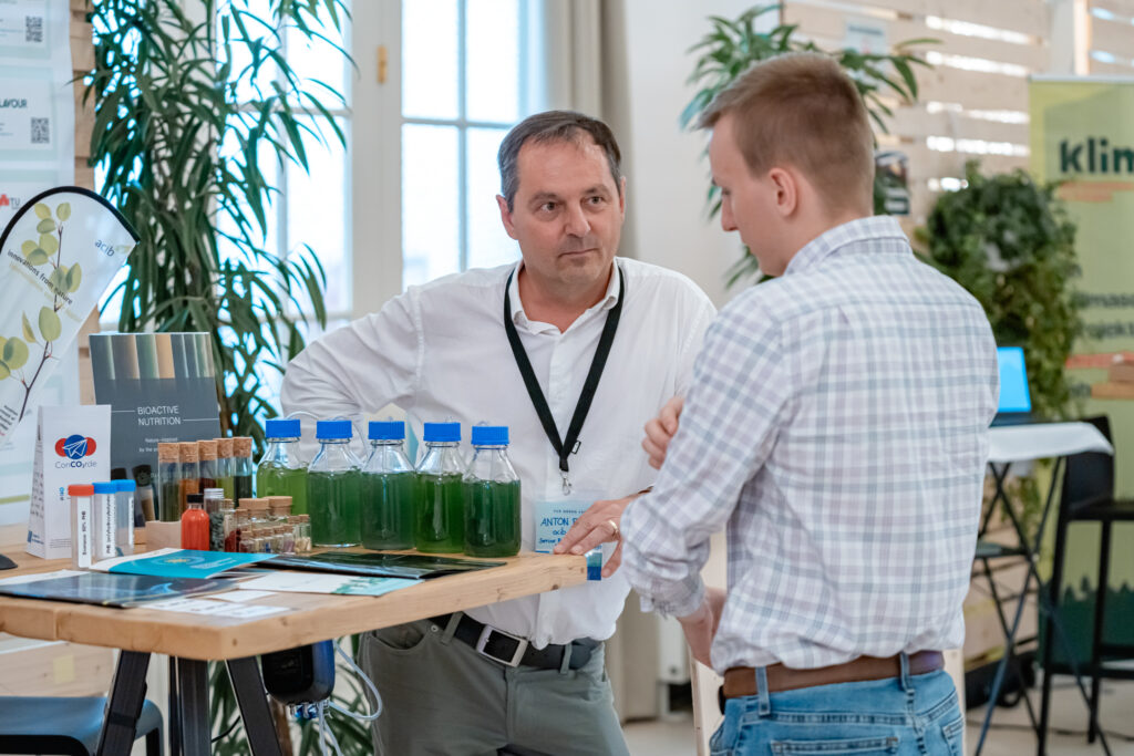 Ein Mann in einem weißen Hemd mit Namensschild und schwarzem Lanyard steht an einem Messestand und spricht mit einem anderen Mann in einem karierten Hemd. Auf dem Tisch stehen mehrere kleine Flaschen mit grünem Flüssigkeitsinhalt, verschiedene Proben in Reagenzgläsern sowie Informationsmaterialien zu biotechnologischen Innovationen. Im Hintergrund sind grüne Pflanzen und Roll-up-Banner zu sehen.