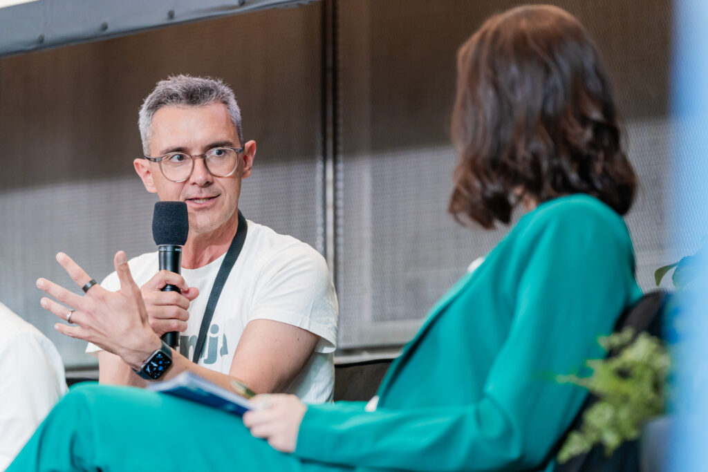 Ein Mann mit grauen Haaren und Brille hält ein Mikrofon und spricht engagiert während einer Podiumsdiskussion. Er trägt ein weißes T-Shirt mit der Aufschrift „klimja“ und eine schwarze Konferenz-Lanyard. Neben ihm sitzt eine Frau im grünen Anzug, die aufmerksam zuhört. Der Hintergrund ist unscharf, mit neutralen Grautönen.