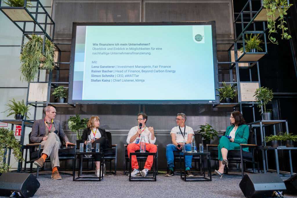Podiumsdiskussion mit fünf Teilnehmenden auf einer Bühne, die von Pflanzen umgeben ist. Im Hintergrund ist eine große Präsentationsfolie zu sehen mit dem Titel „Wie finanziere ich mein Unternehmen?“ sowie den Namen der Diskussionsteilnehmenden: Lena Gansterer, Rainer Bacher, Simon Schmitz und Stefan Kainz. Die Diskussionsteilnehmenden sitzen auf schwarzen Stühlen, während der Mann in der Mitte mit einem Mikrofon spricht.