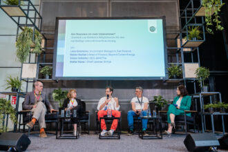 Podiumsdiskussion mit fünf Teilnehmenden auf einer Bühne, die von Pflanzen umgeben ist. Im Hintergrund ist eine große Präsentationsfolie zu sehen mit dem Titel „Wie finanziere ich mein Unternehmen?“ sowie den Namen der Diskussionsteilnehmenden: Lena Gansterer, Rainer Bacher, Simon Schmitz und Stefan Kainz. Die Diskussionsteilnehmenden sitzen auf schwarzen Stühlen, während der Mann in der Mitte mit einem Mikrofon spricht.