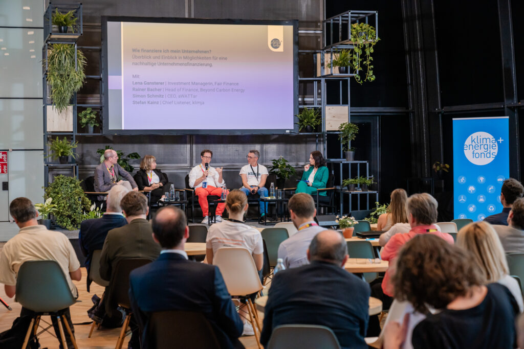 Podiumsdiskussion auf einer Veranstaltung mit fünf Teilnehmenden, die auf einer Bühne vor einem großen Bildschirm sitzen. Auf dem Bildschirm steht der Titel „Wie finanziere ich mein Unternehmen?“ sowie die Namen der Panelisten: Lena Gansterer, Rainer Bacher, Simon Schmitz und Stefan Kainz. Das Publikum sitzt an runden Tischen, während Pflanzen und ein Roll-up mit dem Logo des „Klima + Energie Fonds“ die Bühne dekorieren.