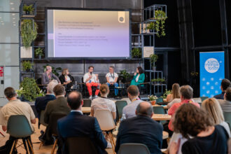 Podiumsdiskussion auf einer Veranstaltung mit fünf Teilnehmenden, die auf einer Bühne vor einem großen Bildschirm sitzen. Auf dem Bildschirm steht der Titel „Wie finanziere ich mein Unternehmen?“ sowie die Namen der Panelisten: Lena Gansterer, Rainer Bacher, Simon Schmitz und Stefan Kainz. Das Publikum sitzt an runden Tischen, während Pflanzen und ein Roll-up mit dem Logo des „Klima + Energie Fonds“ die Bühne dekorieren.