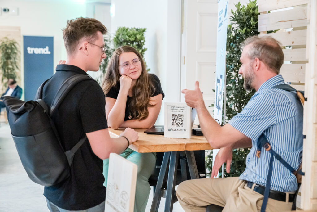 Zwei junge Erwachsene, ein Mann mit schwarzem T-Shirt und Rucksack sowie eine Frau mit langen Haaren und Brille, hören aufmerksam einem Mann mit gestreiftem Hemd und Hosenträgern zu. Sie sitzen an einem hohen Tisch mit einem kleinen Aufsteller, der einen QR-Code und den Text „JETZT FONDS AUF NACHHALTIGKEIT ÜBERPRÜFEN“ von CLEANVEST zeigt. Im Hintergrund sind Pflanzen und ein Banner mit der Aufschrift „trend.“ zu sehen, was auf eine Veranstaltung mit Nachhaltigkeitsthema hinweist.
