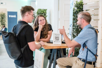 Zwei junge Erwachsene, ein Mann mit schwarzem T-Shirt und Rucksack sowie eine Frau mit langen Haaren und Brille, hören aufmerksam einem Mann mit gestreiftem Hemd und Hosenträgern zu. Sie sitzen an einem hohen Tisch mit einem kleinen Aufsteller, der einen QR-Code und den Text „JETZT FONDS AUF NACHHALTIGKEIT ÜBERPRÜFEN“ von CLEANVEST zeigt. Im Hintergrund sind Pflanzen und ein Banner mit der Aufschrift „trend.“ zu sehen, was auf eine Veranstaltung mit Nachhaltigkeitsthema hinweist.
