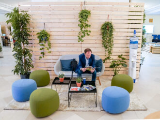 Ein Mann in einem blauen Anzug sitzt auf einem grauen Sofa und liest eine Zeitschrift. Neben ihm steht ein schwarzer Rucksack. Vor ihm befinden sich niedrige Tische mit kleinen Topfpflanzen, Zeitschriften und einem blauen Pappbecher. Um ihn herum sind grüne und blaue Sitzhocker auf einem cremefarbenen Teppich verteilt. Im Hintergrund hängt grüne Dekopflanzen an einer hölzernen Trennwand, rechts steht ein Wasserspender. Die Szene wirkt entspannt und modern, mit einem Fokus auf Natur und Gemütlichkeit.