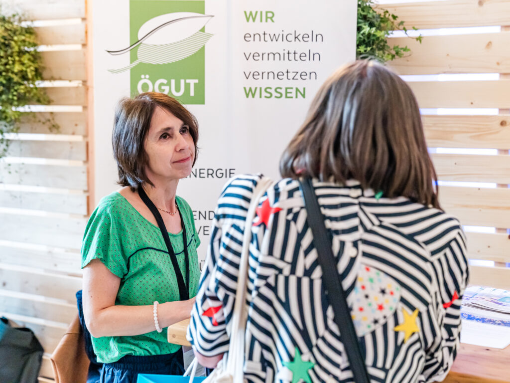 Eine Frau mit braunen Haaren, die ein grünes, gemustertes Oberteil trägt, sitzt an einem Holztisch und spricht mit einer anderen Frau, die von hinten zu sehen ist und ein schwarz-weiß gestreiftes Oberteil mit bunten Sternen trägt. Im Hintergrund steht ein Roll-up-Banner mit dem Logo von 