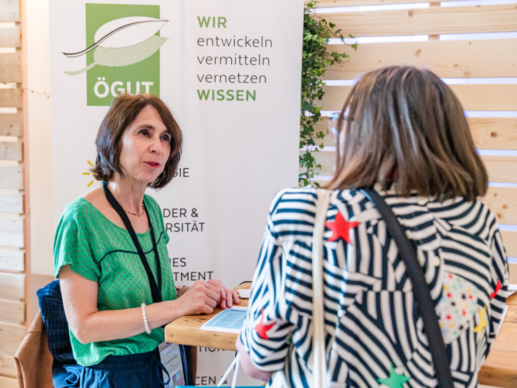 Eine Frau mit braunen Haaren, die ein grünes, gemustertes Oberteil trägt, sitzt an einem Holztisch und spricht mit einer anderen Frau, die von hinten zu sehen ist und ein schwarz-weiß gestreiftes Oberteil mit bunten Sternen trägt. Im Hintergrund steht ein Roll-up-Banner mit dem Logo von 