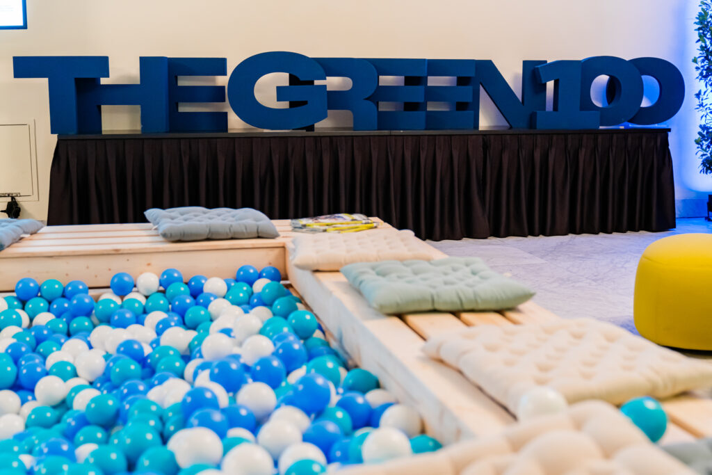 Das Bild zeigt eine kreative Lounge-Ecke auf einem Event, gestaltet mit einer Ballpit-Zone gefüllt mit blauen und weißen Bällen. Umgeben ist die Sitzecke von hellen Holzelementen mit weichen Kissen in Pastelltönen für Komfort. Im Hintergrund steht eine große blaue 3D-Schrift mit dem Schriftzug „THE GREEN 100“, was auf eine nachhaltige oder grüne Veranstaltung hindeutet. Die Atmosphäre wirkt modern, entspannt und einladend.