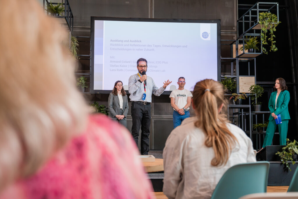 Das Bild zeigt eine Podiumsdiskussion auf einer Bühne mit einer großen Leinwand im Hintergrund, die den Titel „Ausklang und Ausblick“ sowie die Namen der Redner anzeigt. Ein Mann mit Mikrofon spricht, während drei weitere Personen auf der Bühne stehen und zuhören. Eine Frau in einem grünen Hosenanzug hält ein blaues Heft in der Hand. Im Vordergrund sind unscharf zwei Zuschauerinnen zu sehen, die der Diskussion folgen. Die Bühne ist mit modernen Metallregalen und grünen Pflanzen dekoriert, was eine nachhaltige und professionelle Atmosphäre schafft.
